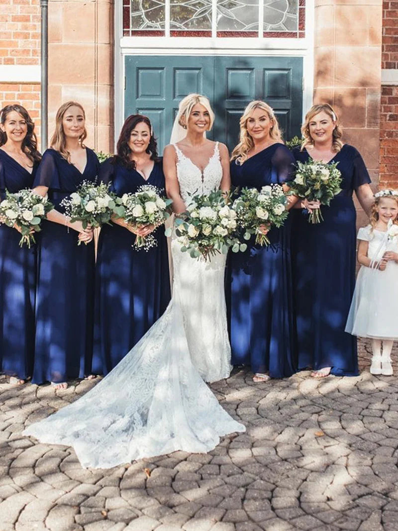Bride in a white dress with bridesmaids in navy blue dresses holding floral bouquets, standing in front of a brick building.