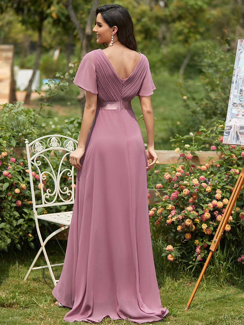 Woman in a long, flowing dress standing in a garden with flowers and a chair.
