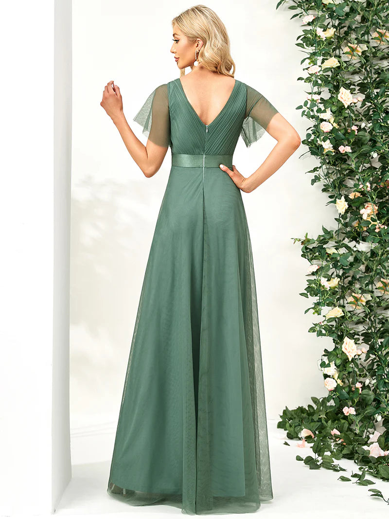Woman wearing a green evening gown with sheer sleeves against a white background with decorative plants.