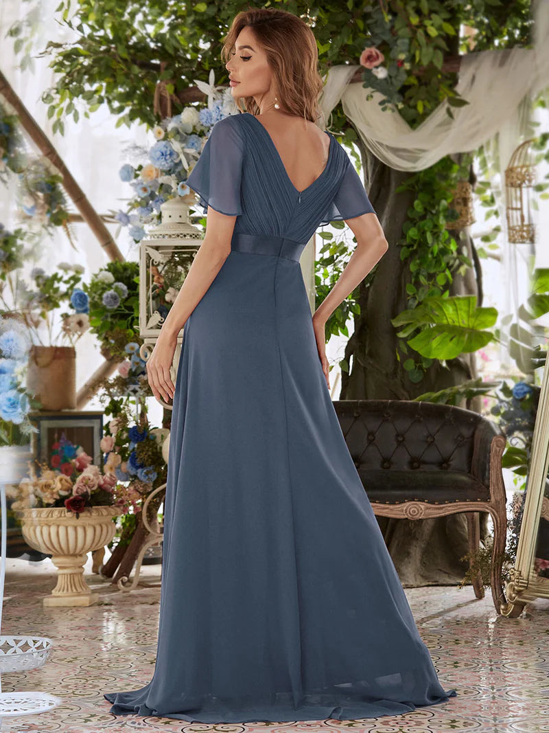 Woman in a dusty blue evening gown standing in an elegant indoor setting with floral decorations.