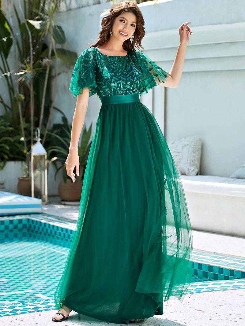 Woman in a dark green dress standing by a poolside with plants and a white wall in the background.