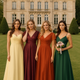 Four bridesmaids standing in front of a grand castle, wearing elegant autumn bridesmaid dresses in butter yellow, burgundy, burnt orange, and dark green.