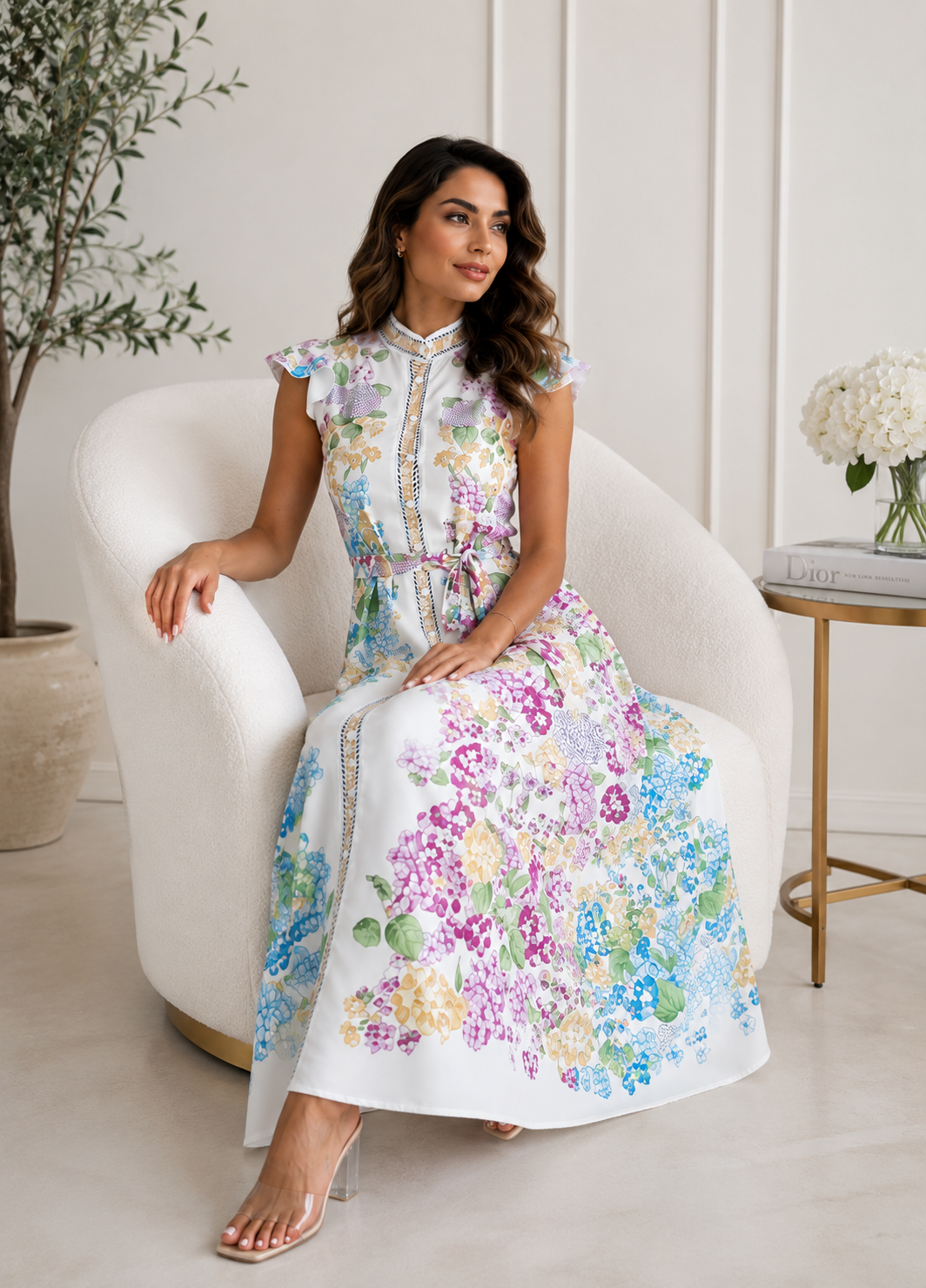 Woman in a floral dress sitting on a white chair in a bright room.