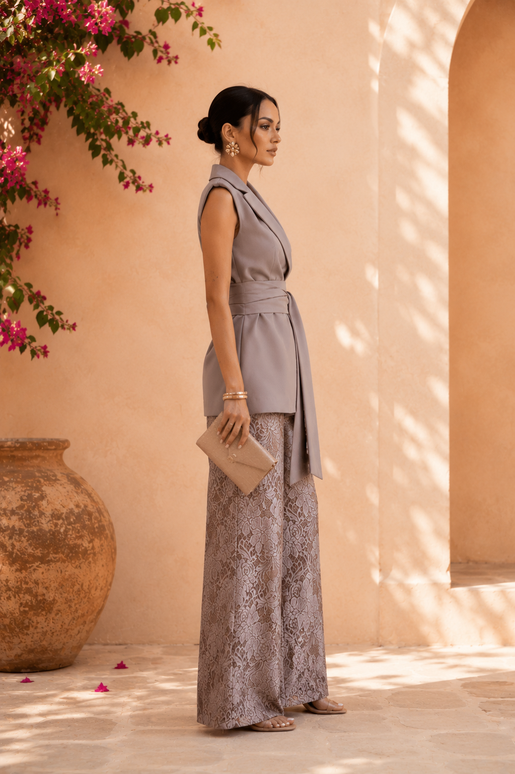 Woman in a gray sleeveless top and patterned pants standing against a beige wall with pink flowers.