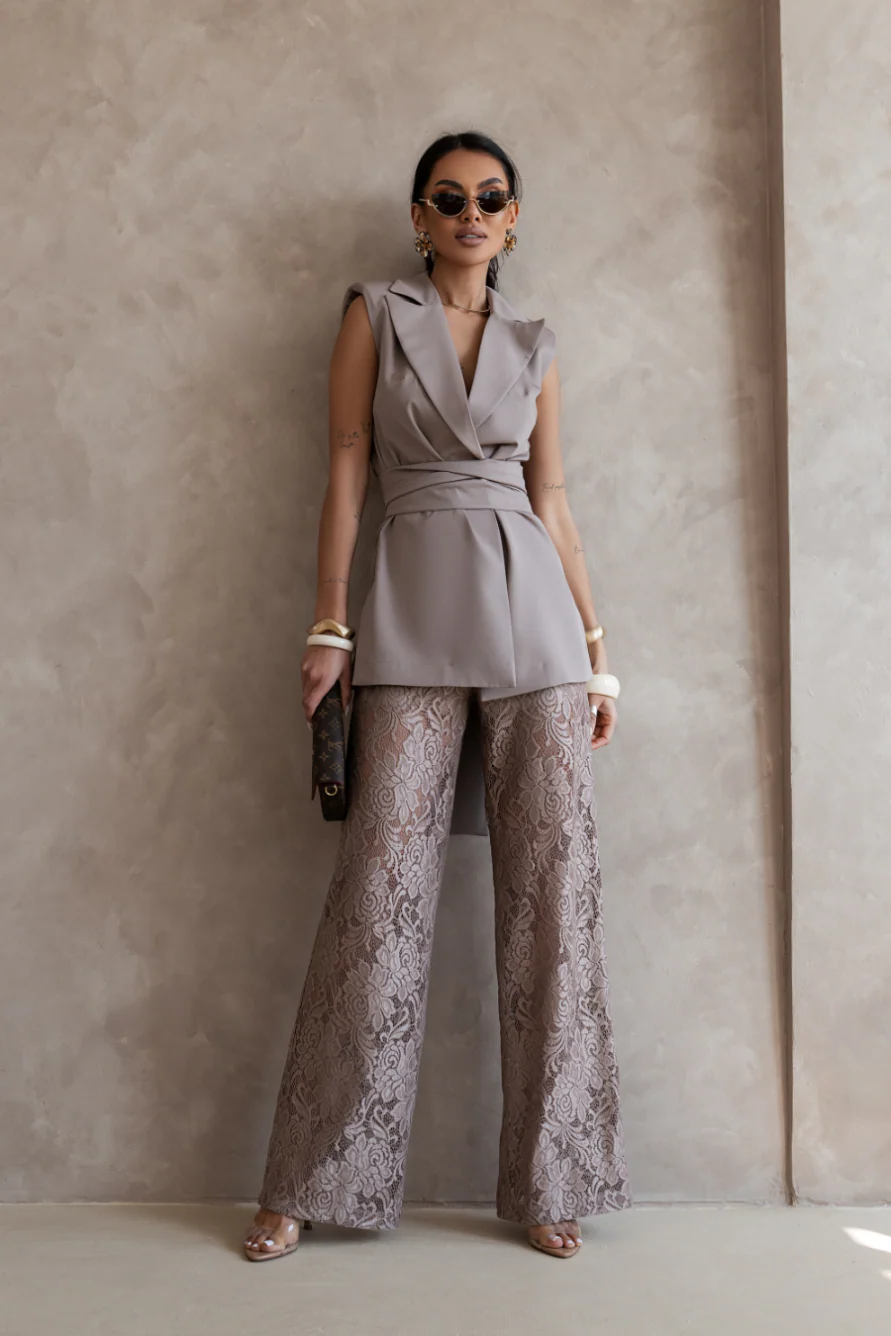 Woman wearing a sleeveless top and lace pants against a plain wall.