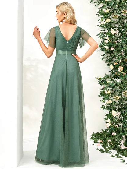 Woman wearing a green evening gown with sheer sleeves against a white background with decorative plants.