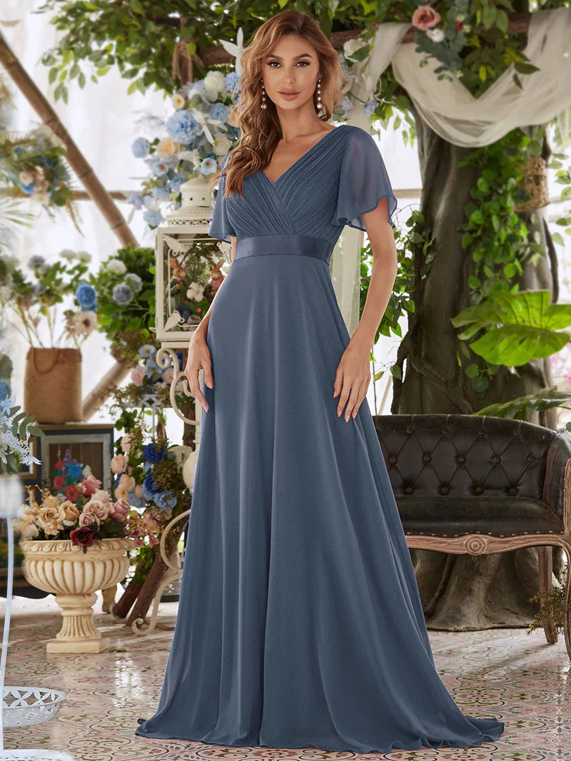 Woman in a Dusty blue gown standing in a decorated indoor setting with flowers and furniture.