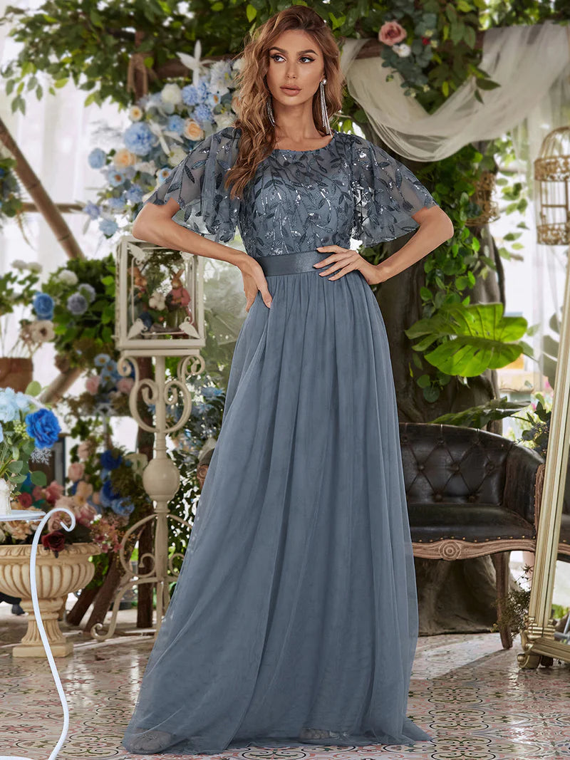 Woman in a Dusty blue evening gown standing in an elegant indoor setting with floral decorations.