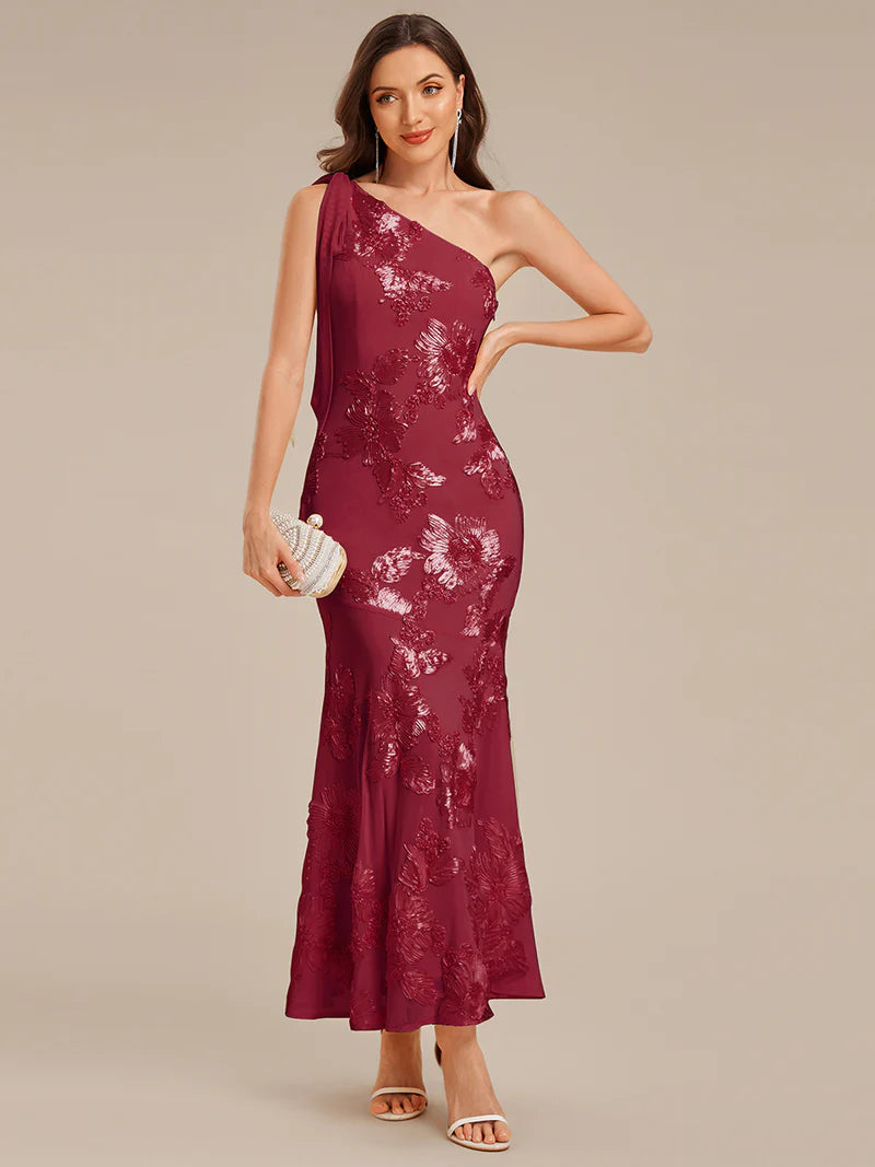 Woman wearing a burgundy one-shoulder dress with floral lace details on a beige background